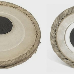 May include: Two Indian tabla drums. The drums have a light tan skin with a dark circular center. The rims are wrapped with a woven rope-like material. The drums are set against a white background.