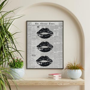 May include: Black and white framed art print featuring three sets of black lips. The background is a newspaper print with the text "The Chicago Times" at the top. The artwork is displayed on a shelf with a plant and books.