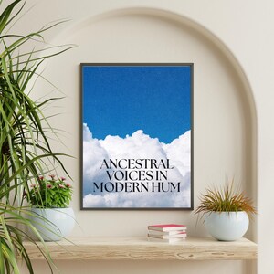 May include: Framed art print featuring a bright blue sky above fluffy white clouds. The text "ANCESTRAL VOICES IN MODERN HUM" is centered below the clouds in a serif font. The artwork is displayed on a wooden shelf with plants and books.