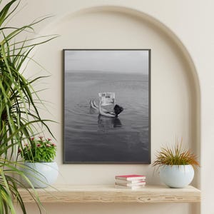 May include: Black and white framed print of a person floating in water while reading a newspaper. The person is wearing a dark swimsuit. The water and sky are a muted gray, creating a serene atmosphere.