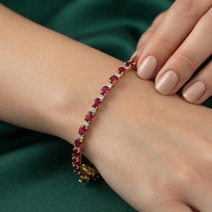 May include: A gold tennis bracelet featuring a series of oval-cut deep red rubies alternating with small, clear, round-cut white stones. The bracelet is secured with a metal clasp and rests on a wrist against a dark green fabric background.