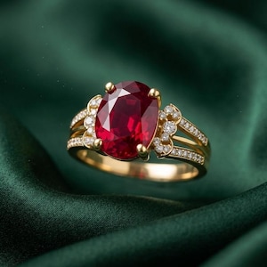 May include: A gold ring featuring a large, oval-cut ruby gemstone as the centerpiece. The ring is adorned with smaller, round diamonds and set against a deep green background. The ring's band has a split design with additional diamond accents.