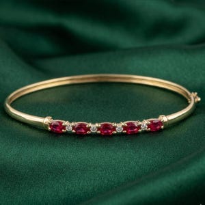 Puede incluir: Una pulsera de oro con una fila de piedras preciosas rojas de corte ovalado y pequeñas piedras blancas. La pulsera está sobre un fondo de satén verde oscuro.