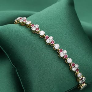 Op de afbeelding: Gouden armband met een bloemmotief. Elke bloem heeft witte en rode edelstenen. De armband wordt getoond op een groene satijnen achtergrond, wat het elegante ontwerp benadrukt.