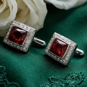 Puede incluir: Un par de gemelos cuadrados con una gran gema roja oscura en el centro, rodeada de piedras claras más pequeñas y brillantes. Los gemelos tienen un engaste y un pasador de metal plateado, sobre un fondo verde oscuro.