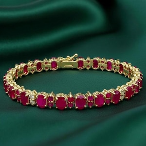 Puede incluir: Una pulsera dorada con piedras preciosas rojas de corte ovalado y pequeñas piedras blancas. La pulsera se muestra sobre un fondo verde oscuro, resaltando su diseño detallado y color.