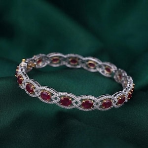 May include: A silver-toned bangle bracelet featuring an intricate, repeating infinity-style link pattern. The design is set with numerous small, clear white stones and accented with oval-shaped, deep red rubies placed at regular intervals.