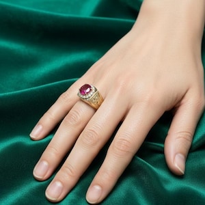 May include: A gold ring featuring a large, oval-cut ruby gemstone surrounded by small, clear accent stones. The ring has an ornate, detailed design on the band. The ring is displayed on a hand against a green background.