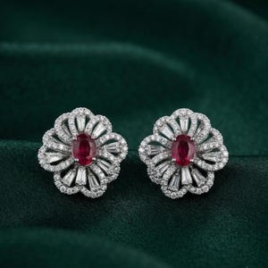 May include: A pair of silver-toned flower-shaped earrings featuring a central oval-cut red gemstone surrounded by a halo of small round white stones and radiating baguette-cut white stones arranged in a petal-like pattern.