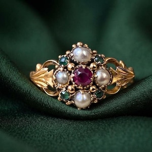 May include: A gold ring featuring a central round red gemstone surrounded by four white pearls and four small green gemstones. The band is crafted with intricate leaf-like metalwork on both sides, creating a detailed and ornate appearance.