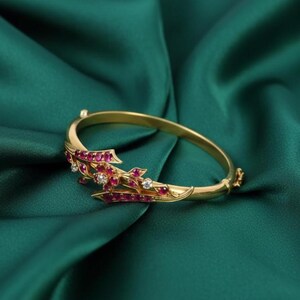 May include: A gold bracelet with a floral design, set with red gemstones and small white diamonds. The bracelet is displayed on a deep green satin fabric, accentuating its intricate details and luxurious appearance.