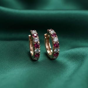 May include: A pair of gold hoop earrings, each adorned with alternating red and clear gemstones. The earrings are set against a dark green, silky background, highlighting the sparkle of the jewelry.