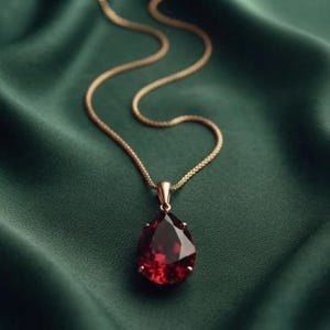 May include: A necklace featuring a teardrop-shaped deep red gemstone pendant suspended from a delicate gold-toned chain. The pendant is secured by a small gold-toned metal bail, and the faceted stone reflects light with a dark crimson hue.