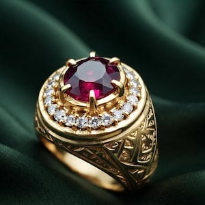 May include: A gold ring featuring a large, round, deep red gemstone set in the center, surrounded by a halo of small, clear, sparkling stones. The gold band displays an intricate, textured, geometric pattern along its outer surface.