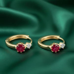 以下が含まれることがあります： 鮮やかな赤い宝石と小さなクリアストーンが付いた、2つのゴールドフープイヤリング。イヤリングは、深緑色のテクスチャード加工された背景に配置され、コントラストを生み出しています。