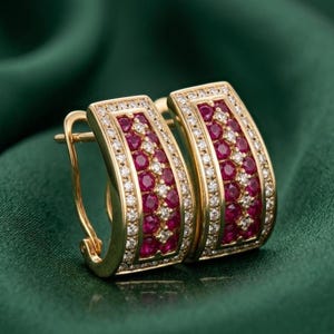 May include: A pair of gold-toned hoop earrings. Each earring features a rectangular design with a row of small, round, red gemstones in the center, surrounded by a row of small, clear gemstones. The earrings are set against a green background.