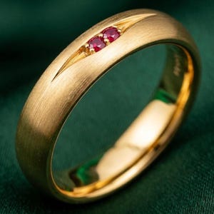 Puede incluir: Un anillo de banda de oro cepillado con un acabado mate y liso en la superficie exterior. El anillo está decorado con dos pequeños rubíes rojos redondos y vibrantes, engastados uno al lado del otro en un canal estrecho y alargado.