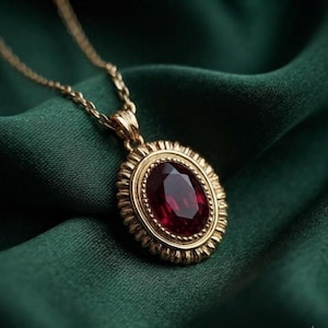 May include: A gold pendant necklace featuring an oval-shaped, deep red faceted gemstone set within a textured, scalloped gold frame. The pendant hangs from a delicate gold chain, showcasing a classic design with a polished metal finish.