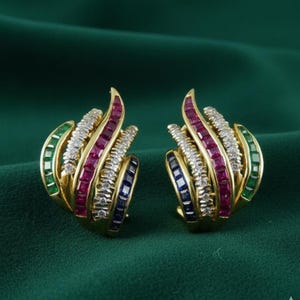 Puede incluir: Un par de pendientes dorados con un diseño curvo. Cada pendiente presenta filas de piedras preciosas verdes, rojas, blancas y azules. Los pendientes están sobre un fondo verde oscuro.