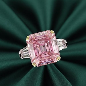 May include: A large, square-cut pink gemstone ring with a gold setting and two baguette-cut side stones. The ring is set against a dark green background, highlighting the brilliance of the pink stone.