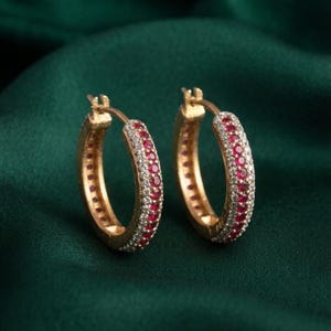 May include: A pair of gold hoop earrings featuring a central row of round red rubies flanked by two rows of small white diamonds. The earrings have a hinged closure mechanism and are designed with a circular shape, showcasing a polished gold finish.
