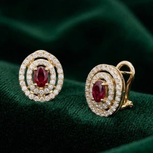 May include: A pair of gold earrings featuring a central oval-cut red ruby surrounded by two concentric halos of small round white diamonds. The earrings have a secure post and hinge back mechanism for attachment to the earlobe.