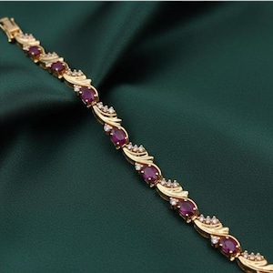 May include: A gold-coloured bracelet with oval, deep red gemstones and small, sparkling white stones. The bracelet has an ornate design and a secure clasp. The jewellery is displayed against a dark green background.