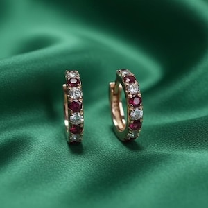 Może przedstawiać: Para złotych kolczyków koła, każdy ozdobiony naprzemiennie czerwonymi i przezroczystymi kamieniami szlachetnymi. Kolczyki są umieszczone na głębokim, zielonym, jedwabistym tle, podkreślając blask i design biżuterii.