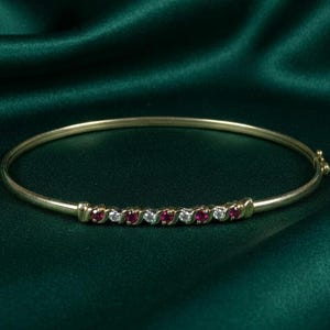 Op de afbeelding: Een gouden armband met afwisselend rode en heldere edelstenen. De armband is geplaatst tegen een donkergroene achtergrond, die de glans van de edelstenen benadrukt. Een klassiek sieraad.