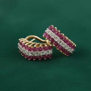 Pode incluir: Um par de brincos dourados com um design retangular. Os brincos são adornados com fileiras de pequenas pedras preciosas redondas, vermelho escuro, e uma fileira central de pedras preciosas menores e transparentes. Os brincos estão em um fundo verde escuro.