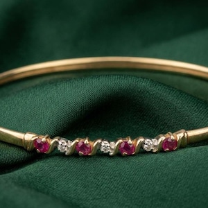 Könnte beinhalten: Ein goldener Armreif mit abwechselnd roten Edelsteinen und kleinen weißen Edelsteinen. Der Armreif liegt auf einem dunkelgrünen Stoffhintergrund.
