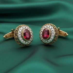 May include: A pair of gold cufflinks with oval-shaped ruby-red center stones, surrounded by small white gemstones and a decorative gold filigree border. The cufflinks are set against a dark green background.