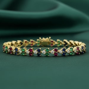 May include: A gold-toned bracelet with a repeating pattern of colorful gemstones. The gemstones are arranged in a chevron design, with red, blue, green, and white stones. The bracelet is set against a dark green background.