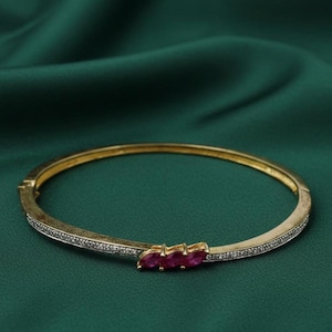 Puede incluir: Un brazalete dorado con tres piedras preciosas ovaladas de color rojo intenso. El brazalete tiene un cierre de bisagra y está engastado con pequeñas piedras transparentes a lo largo de la banda. El fondo es una tela verde oscuro texturizada.