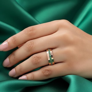 Könnte beinhalten: Ein roségoldener Ring mit einer Reihe kleiner, leuchtend grüner Edelsteine. Der Ring ist vor dem Hintergrund eines tief smaragdgrünen Stoffes platziert, was einen auffälligen Kontrast erzeugt. Das Design des Rings ist schlicht und elegant.