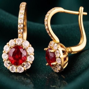 May include: A pair of gold hoop earrings featuring a central round-cut red gemstone surrounded by a halo of small, clear, round-cut white stones. The hoop mechanism is also encrusted with a row of clear white stones, creating a cohesive design.