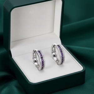 May include: Two silver hoop earrings, each featuring a line of purple gemstones and a line of clear stones. They are presented in a dark green jewellery box with a white interior, against a dark green satin backdrop.