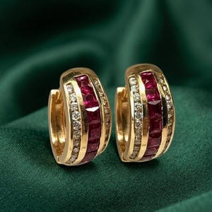 May include: A pair of gold hoop earrings featuring a central row of square-cut deep red rubies flanked by two rows of small, clear round-cut diamonds. The earrings have a hinged closure mechanism and a polished metallic finish throughout.