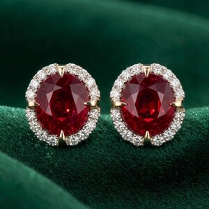 May include: A pair of oval-shaped ruby earrings. Each earring features a large, deep red gemstone surrounded by a halo of small, sparkling white diamonds. The setting is gold-toned, and the earrings are displayed on a dark green velvet surface.