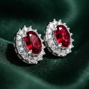 May include: A pair of stud earrings featuring oval-cut deep red rubies as the center stones. Each ruby is surrounded by a halo of small, round, brilliant-cut white diamonds set in a polished silver-toned metal frame with a secure prong setting.