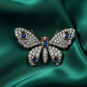 May include: A butterfly-shaped brooch with a gold frame, encrusted with small, clear gemstones. The wings feature four square, deep blue gemstones. The body has a central blue gemstone and a small red gemstone. The brooch rests on a green satin fabric.