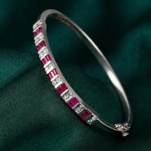 May include: A silver-toned hinged bangle bracelet featuring a row of alternating rectangular red rubies and small clear round diamonds. The metal band has a polished finish and a secure clasp mechanism visible at the bottom of the circular frame.