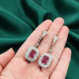 Puede incluir: Un par de pendientes colgantes vintage con gemas rectangulares de color rosa intenso, rodeadas por un halo de pequeñas piedras claras. Los pendientes tienen un engaste dorado, sobre un fondo verde.