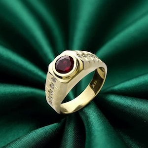 May include: A gold ring with a large, round, deep red gemstone at the center. The ring has a rectangular face and is accented with small, clear gemstones. The band is marked with "14K".
