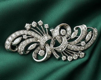 May include: A silver-toned brooch with a floral design, encrusted with numerous clear, sparkling gemstones. The brooch features curved lines, leaf-shaped accents, and a symmetrical arrangement, set against a dark green fabric.