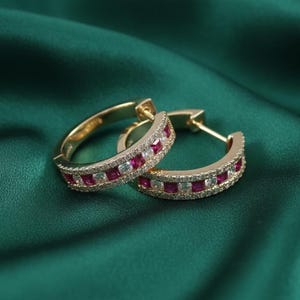 Puede incluir: Un par de pendientes de aro dorados con gemas rojas y claras alternas. Los pendientes se colocan sobre un fondo verde oscuro, resaltando el brillo de las joyas. Las gemas son de talla cuadrada.