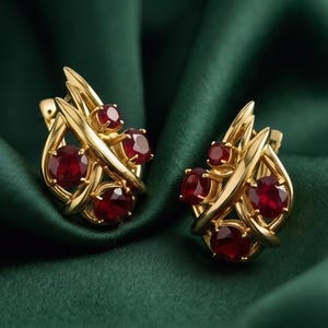 Könnte beinhalten: Ein Paar goldfarbene Ohrringe mit roten Edelsteinen. Das Design weist geschwungene Linien auf und ist vor einem dunkelgrünen Hintergrund platziert. Ein elegantes Schmuckstück.