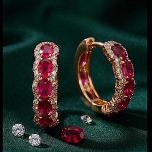 Könnte beinhalten: Ein Paar goldene Creolen mit einer Reihe oval geschliffener roter Rubine, die von kleinen weißen Diamanten umrandet sind. Ein Ohrring steht aufrecht, der andere ist geöffnet. Zwei lose weiße Diamanten und ein loser ovaler Rubin liegen daneben.