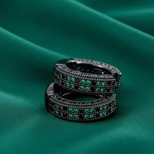 May include: Two black hoop earrings with rows of small, sparkling green gemstones. The earrings are set against a dark green, silky background, creating a striking contrast. The jewelry has a modern, elegant design.
