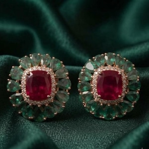 May include: A pair of stud earrings featuring a central cushion-cut deep red gemstone surrounded by a halo of small white diamonds and an outer border of pear-shaped green gemstones arranged in a floral pattern set in gold metal.
